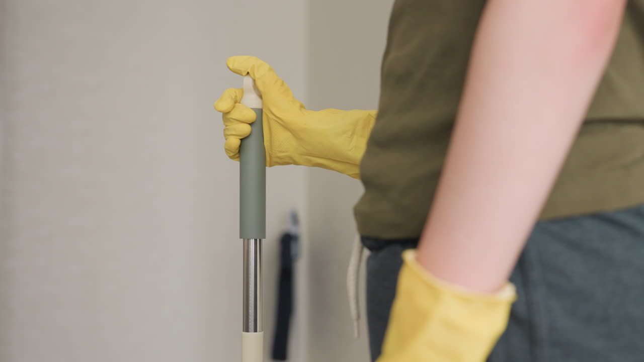 Close up partial view of maid in casual clothes wearing yellow gloves holding mop, placing one hand over the other, focus on protective gloves and cleaning posture during household work indoors