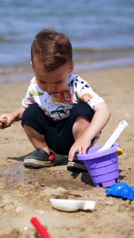 adorable toddler boy plays with toys at the seashore. kid pours some water from his bucket on the sand. vertical video