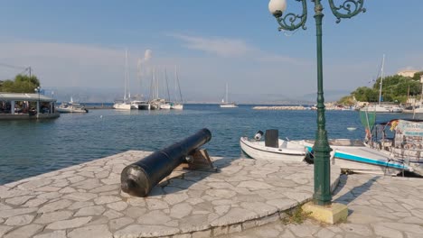 Cañón-De-Guerra-En-El-Pintoresco-Paseo-Marítimo-De-Kassiopi,-Barcos-De-Pesca-Amarrados-Durante-El-Verano,-Isla-De-Corfú