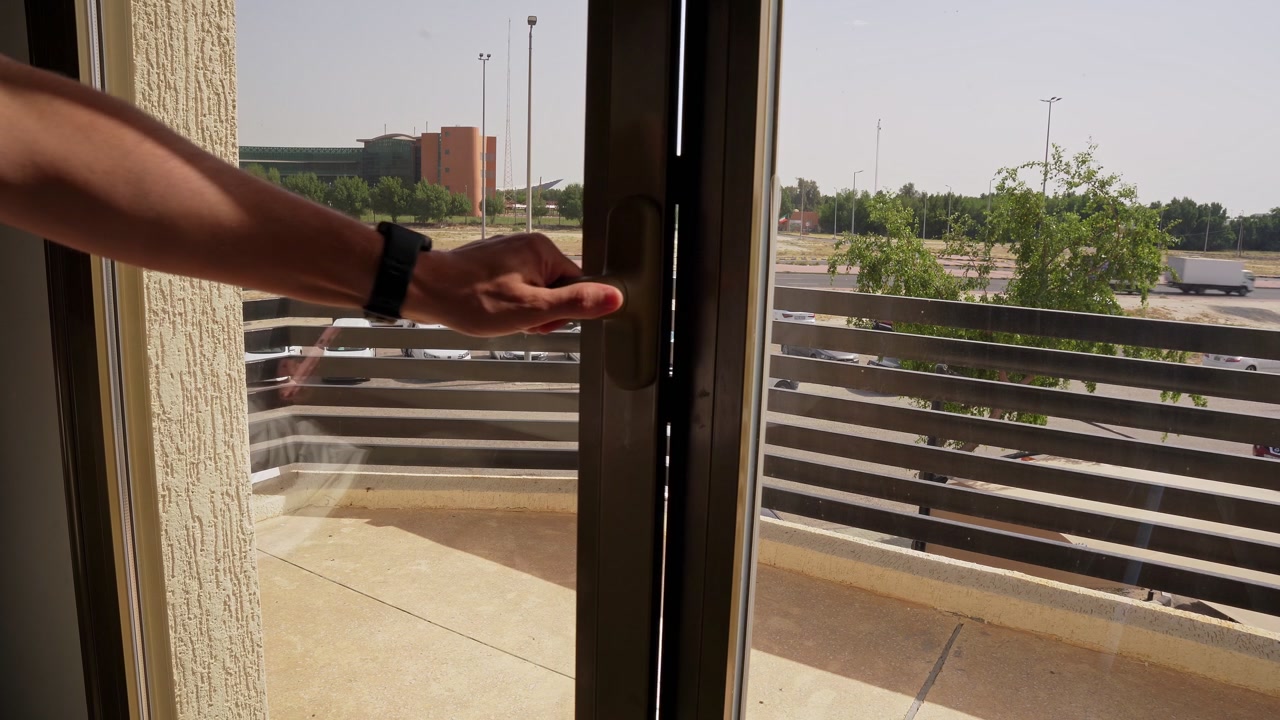 Primer plano de la mano de un hombre abriendo y cerrando una puerta corredera de cristal de balcón. Con vistas a una soleada vista urbana de la ciudad con edificios residenciales y árboles verdes.