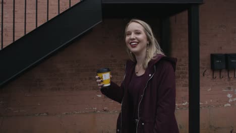 slow motion of a young woman smiling and walking as she raises her coffee cup towards the camera with a red brick wall and staircase in the background