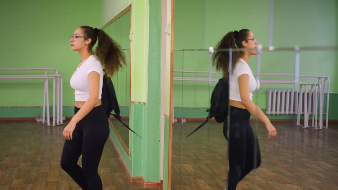 Lady in casual wear with school bag walks out and closes door, mirror reflecting her body along with iron bars in bright indoor room featuring polished wooden floor and green painted walls