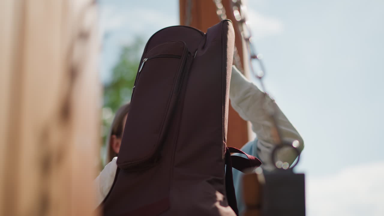 white guitarist packing guitar case outdoors on wooden swing, sunlight filtering through trees, careful hands fastening straps, padded case lifted against sky, contemplative songwriter preparing
