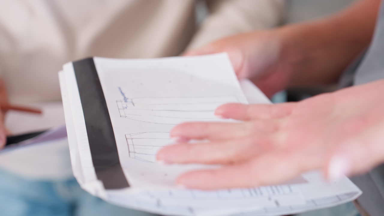 Closeup view of female hands pointing at trouser design sketch drawn on paper sheet during fashion project discussion teamwork clothing development planning creative collaboration session indoors
