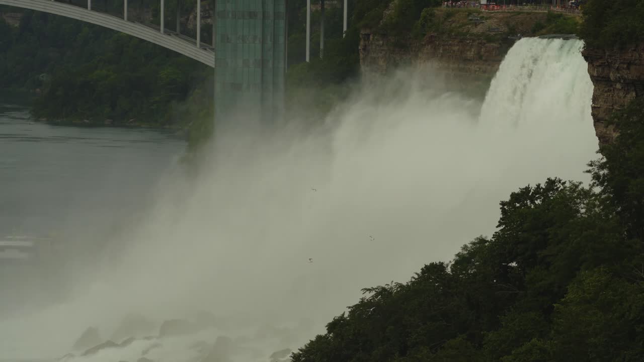 Massive mist rises as powerful waterfalls crash below a scenic bridge at Niagara Falls, surrounded by lush green trees and dramatic cliffs
