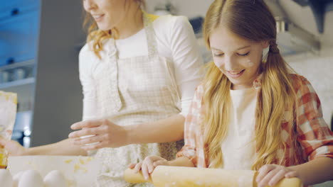 Mother-and-daughter-making-a-daugh-for-the-cookies.-Mom-pouring-a-flour-while-a-daughter-kneading-it-on-the-kitchen-table-in-the-evening.-Portrait-shot.-Inside