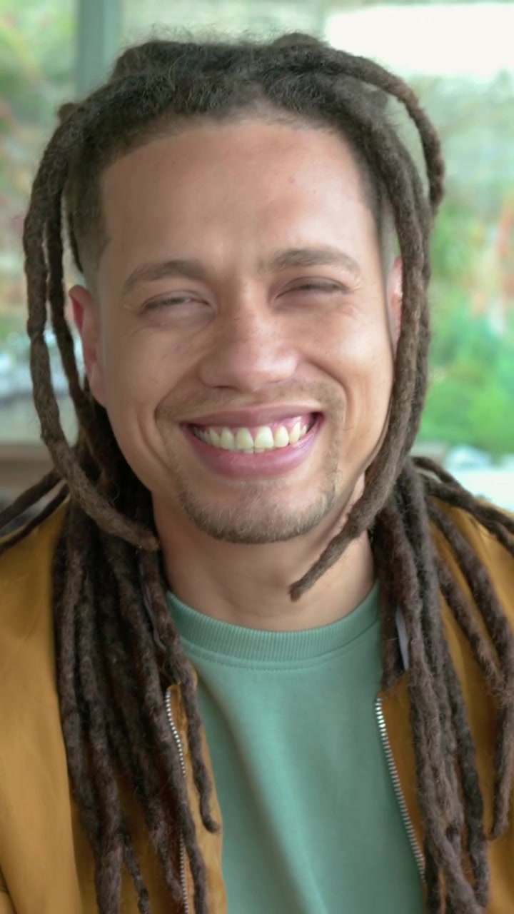 Vertikal video: Smilende mann med dreadlocks snur seg mot kamera på sofaen når han hører latter, jakke synlig.