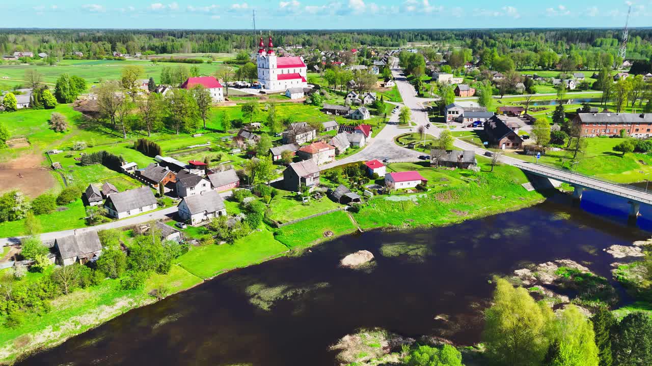Lush green Lithuanian village with houses, roads, and a river under a sunny sky