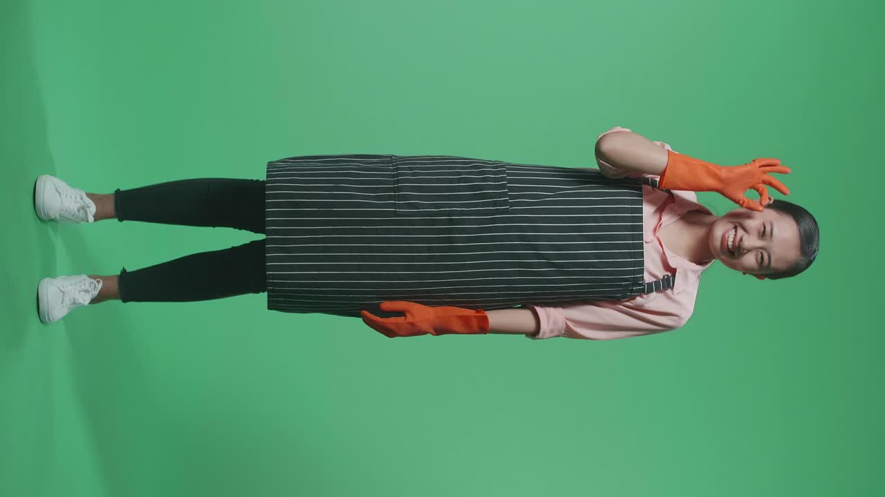 녹색 스크린 스튜디오에 서 있는 동안  제스처와 미소를 짓고 있는 아프론을 입은 아시아 여성 집주인의 몸이 가득 차 있습니다.