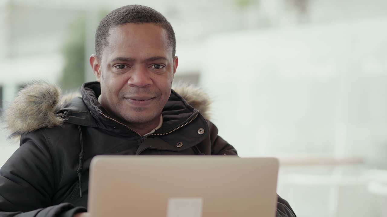 hombre afroamericano de mediana edad con chaqueta negra y capucha de piel trabajando en una computadora portátil afuera