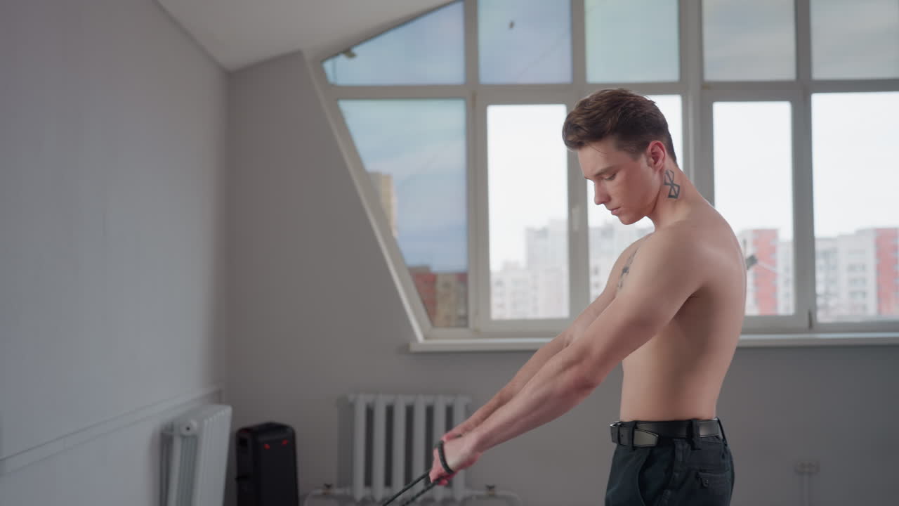 young man with chest tattoo wraps rope around hand and pulls with focused effort in bright studio by window, muscles flexing during resistance training