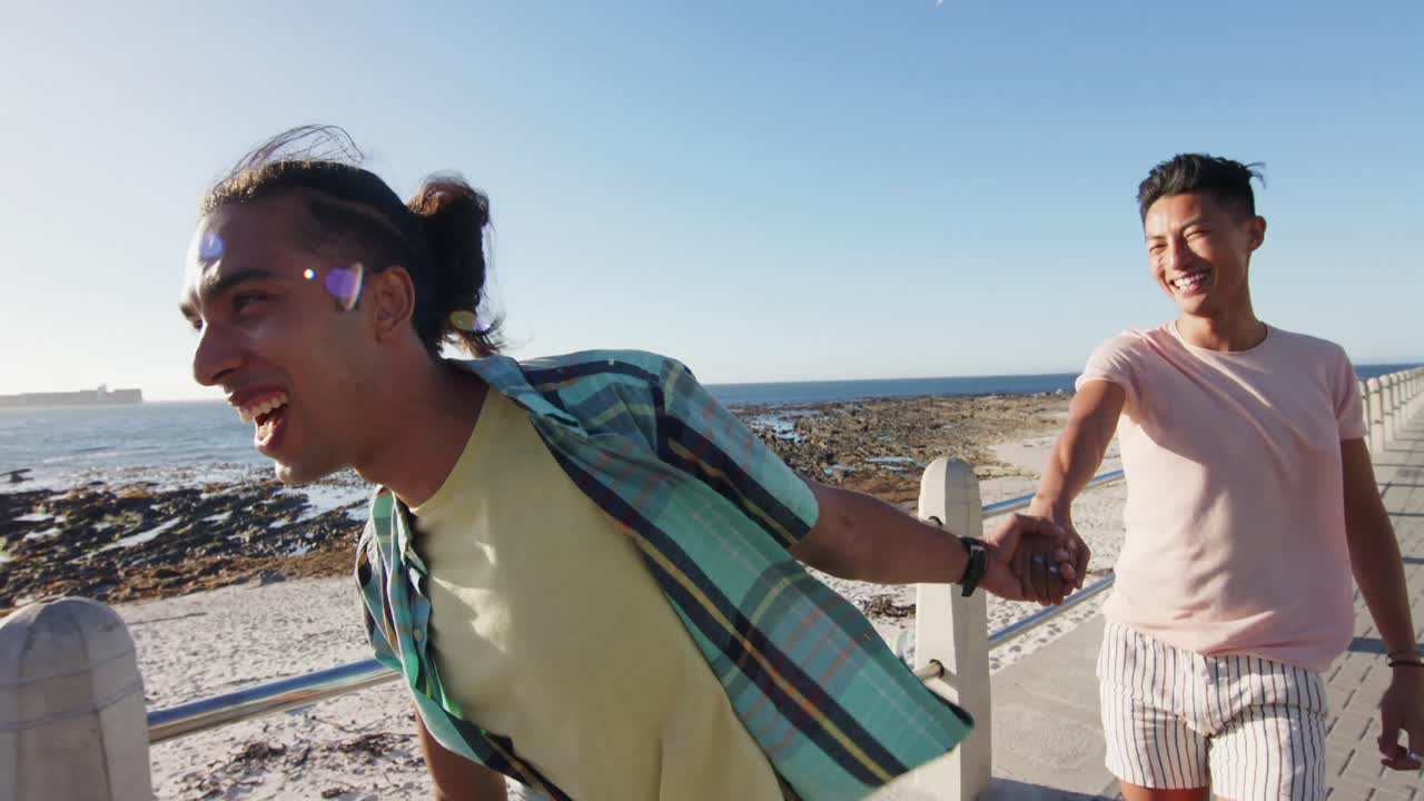 해변의 산책로에서 손을 잡고  ⁇ 고 있는 행복한 다양한 동성애 남성 커플, 느린 동작