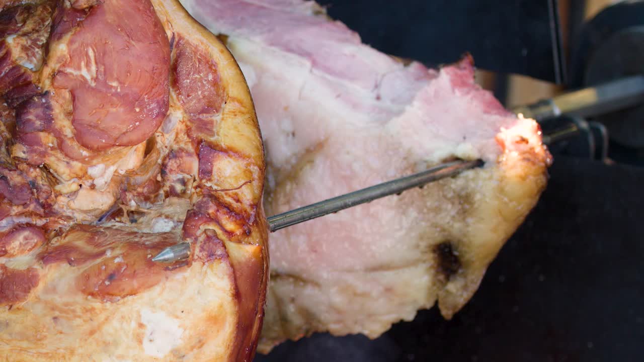 Gloved hand turns pork ham on spit over open flames at outdoor street food market