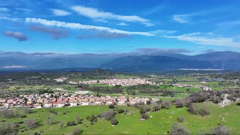 vuelo lateral con un dron en el valle donde vemos una ciudad rural, una zona verde de pasto con ganado y en el fondo el sistema de montañas peninsular central con un cielo azul con nubes