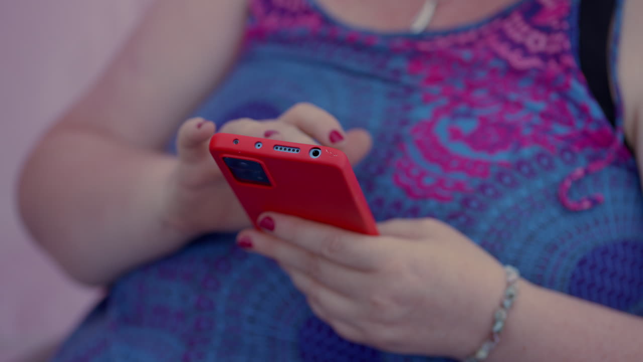 un primer plano de una mujer gorda desplazándose y jugando con su teléfono inteligente rojo mientras está sentada, con un colorido vestido rosa azul y esmalte de uñas rojo en cámara lenta