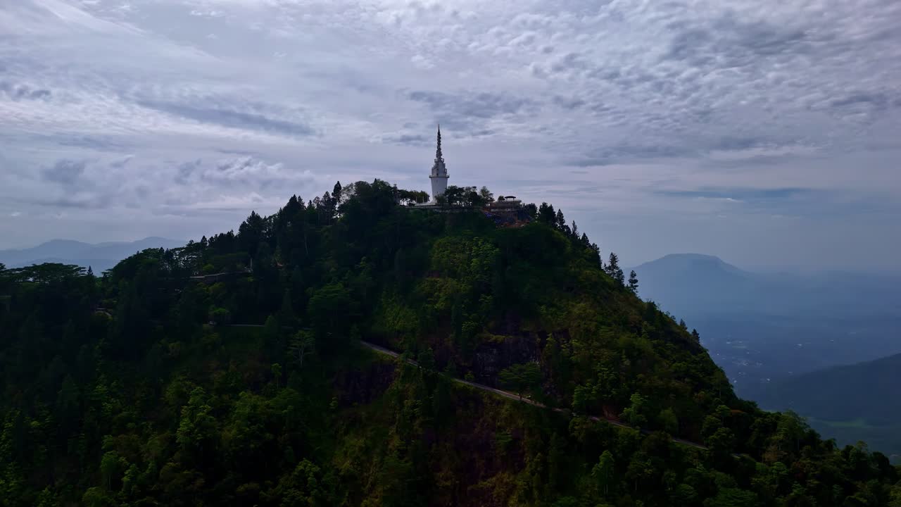 A tall slender tower stands atop a green mountain, overlooking rolling hills under a cloudy sky