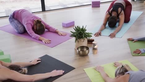 Diverse-women-practicing-child-pose-around-plant-and-singing-bowl-in-yoga-studio