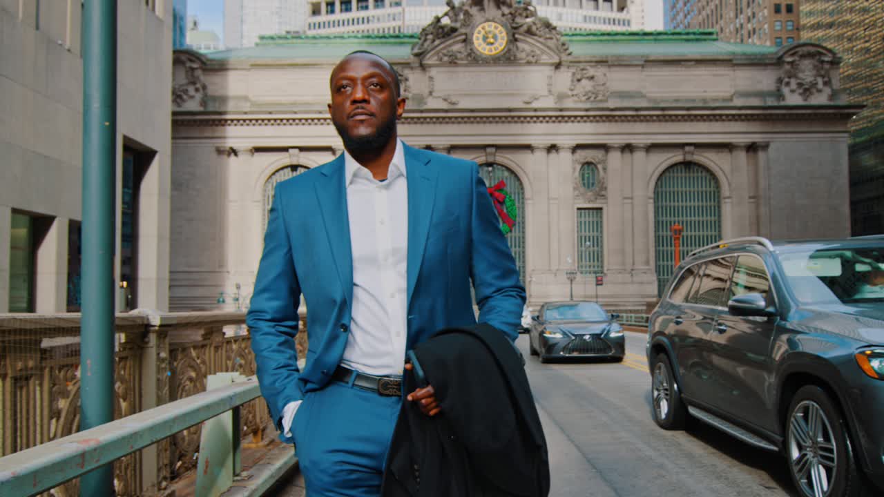 New York, USA, 15 aprile 2026: Uomo d'affari che cammina su un ponte con il Grand Central Terminal sullo sfondo. Vista frontale di un uomo di colore in abito che cammina verso la telecamera vicino al Grand Central Terminal a New York