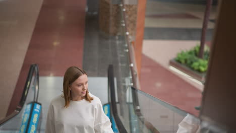 lady riding moving escalator with one hand on rail, wearing casual white shirt, looking down, with blurred background showcasing modern shopping mall interior and plants in the surroundings