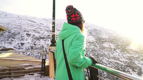 mujer con una chaqueta verde y un sombrero caminando hacia un mirador con paisaje nevado en un día soleado