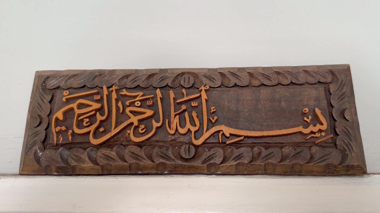 A wooden plaque featuring hand-carved Arabic calligraphy of the an arab phrase, framed by an ornate braided style border. The raised lettering is finished in gold tone against brown wood.