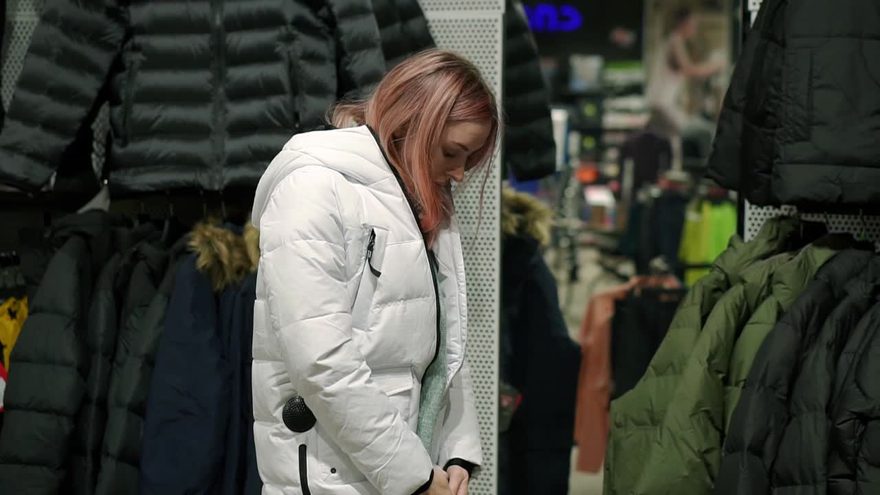 Woman is taking a new down jacket from a rack to fit it, look at the mirror