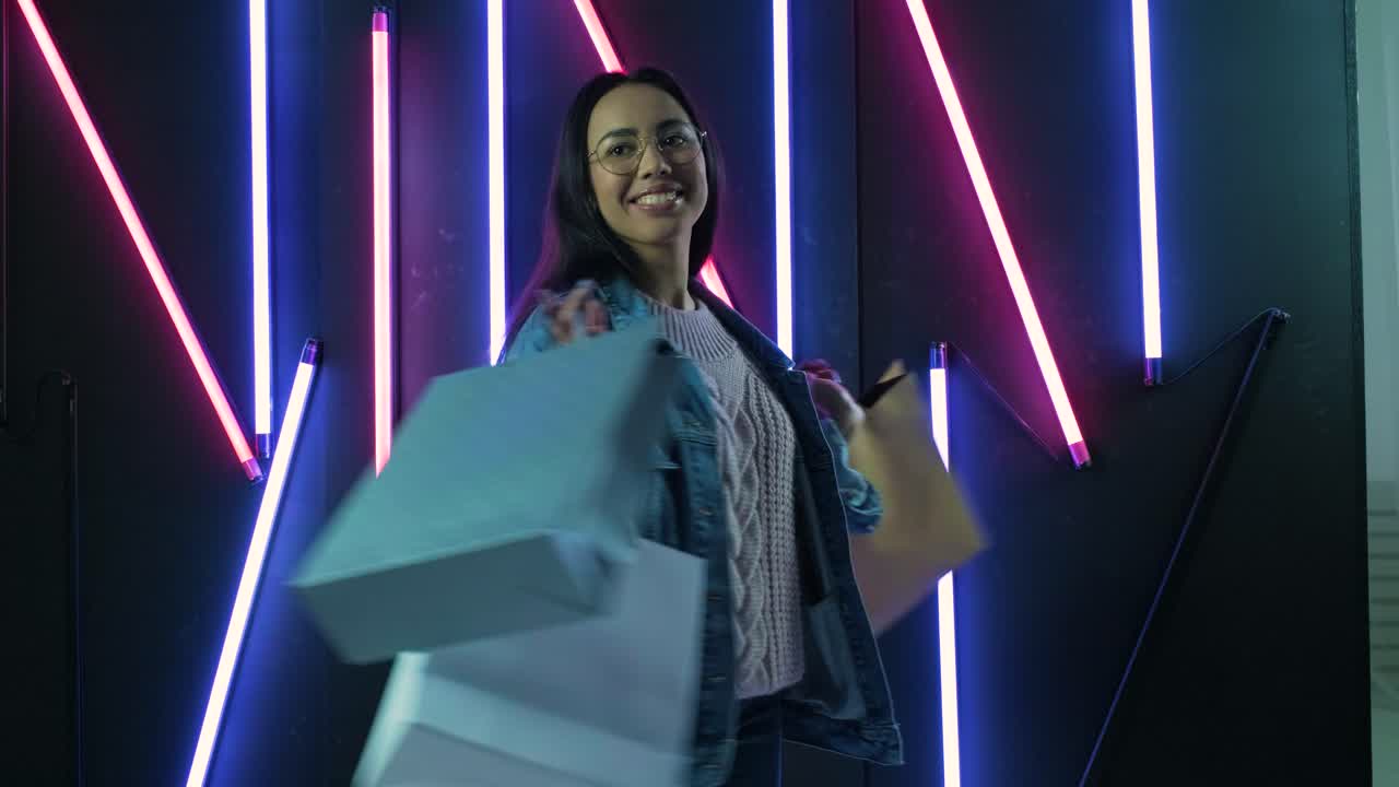 joven con chaqueta de vaqueros azul con bolsas de compras en el fondo de lámparas de neón.