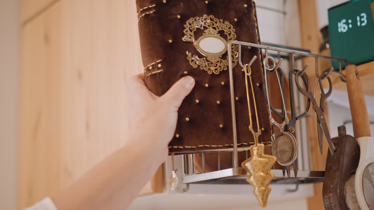 Diario in velluto e gioielli di famiglia, primo piano di una mano che prende un libro decorato da uno scaffale di metallo accanto a un mobile di legno, pendolo in ottone e catene a strati drappeggiati sulla copertina, luce ambientale calda, atmosfera rustica e accogliente.