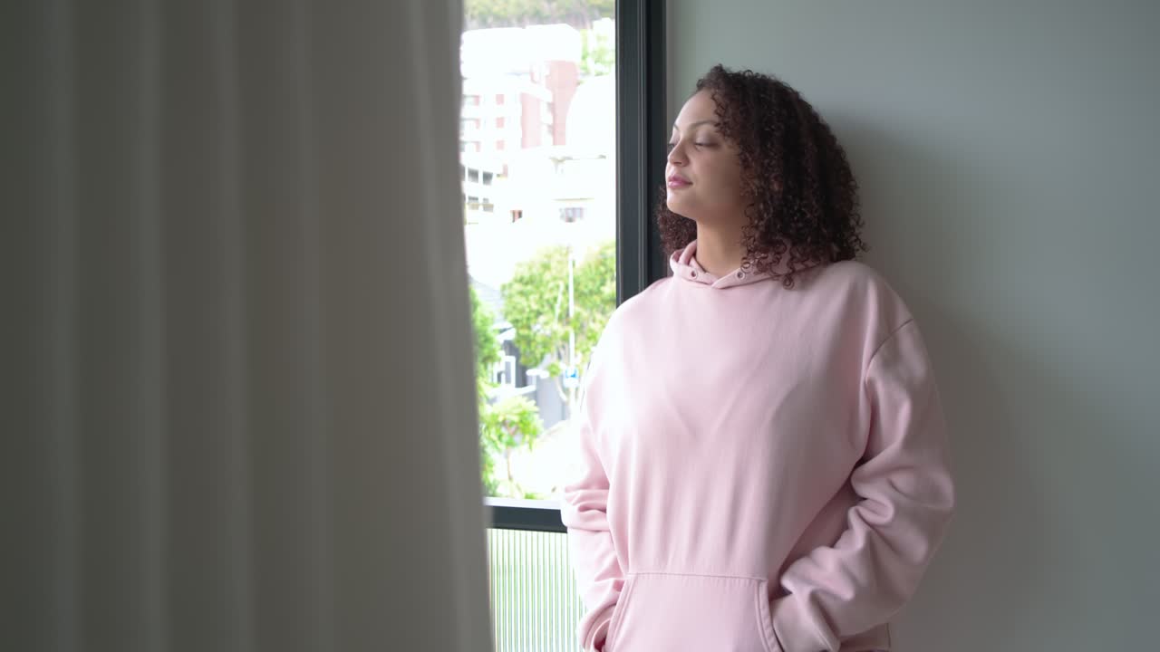Woman leaning in apartment by window in pink hoodie, noticing street view, reflecting, smiling