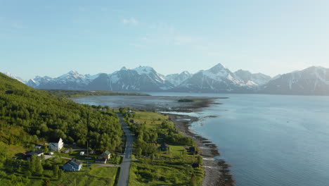 Vista-Aérea-De-Casas-Y-Una-Carretera-Costera,-El-Mar-ártico-Y-El-Fiordo-Nevado-De-Montaña-En-El-Fondo,-Soleado,-Día-De-Verano,-En-Rotsund,-Troms,-Nordland,-Noruega---Levantamiento,-Disparo-De-Drones