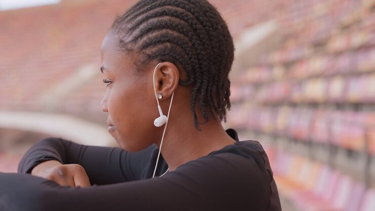 young woman in stadium leaning on railing with head down, earphones in, exhausted and reflective closeup of athlete catching breath, empty bleachers background, quiet healing mood