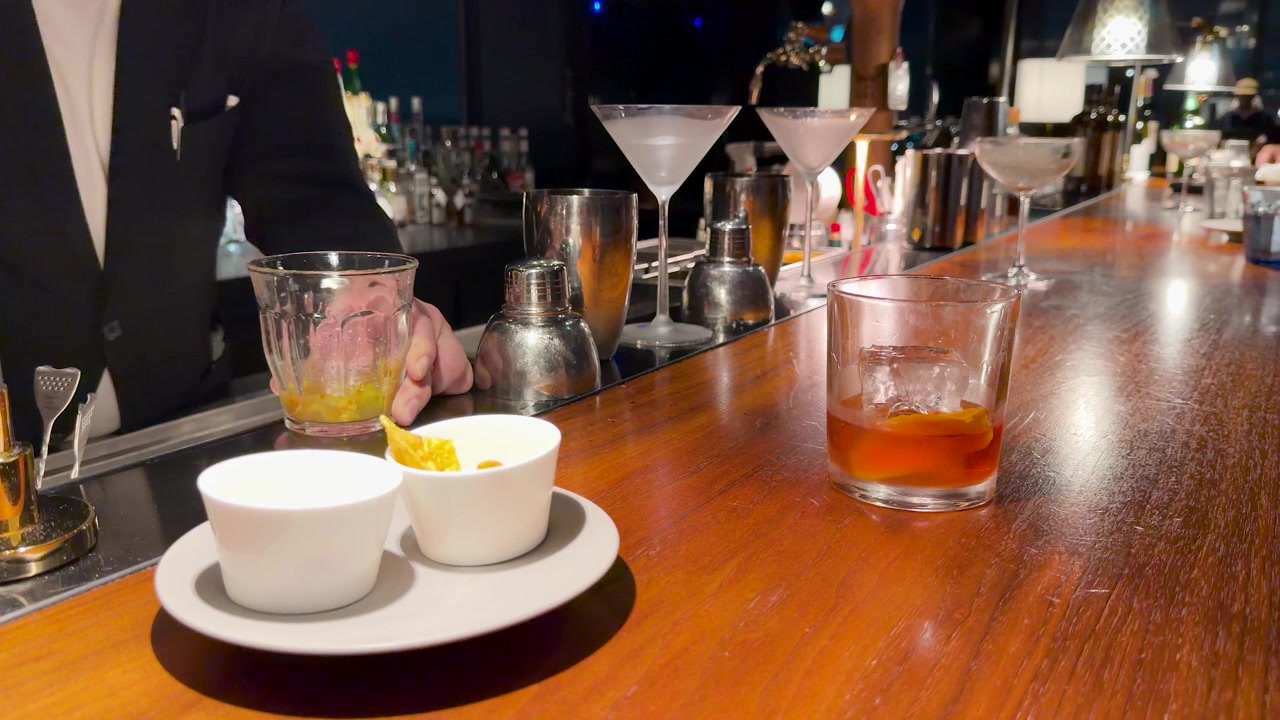 Một người pha chế mặc vest đang dầm các nguyên liệu tươi tại quầy bar gỗ bóng loáng.