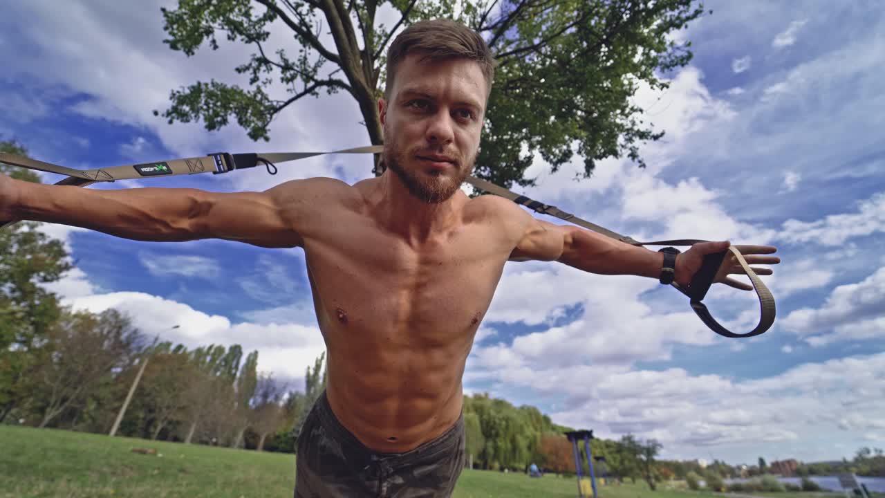 Shirtless sportsman exercising with trx in nature. Muscular athlete is training his strong body near the tree under blue sky in summer. View from below.