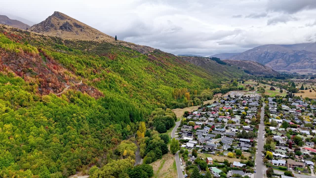 Aerial drone follows Tobins Track with colorful autumn trees on the hillside and Arrowtown appearing to the right under cloudy skies