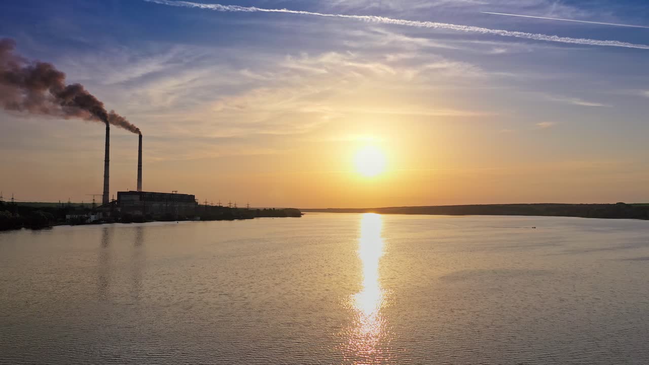 Drone landscape of sunset view on the river surface. Beautiful evening river with sun reflection. Industrial pipes with smoke from the plant near the water. Camera moves bottom up.