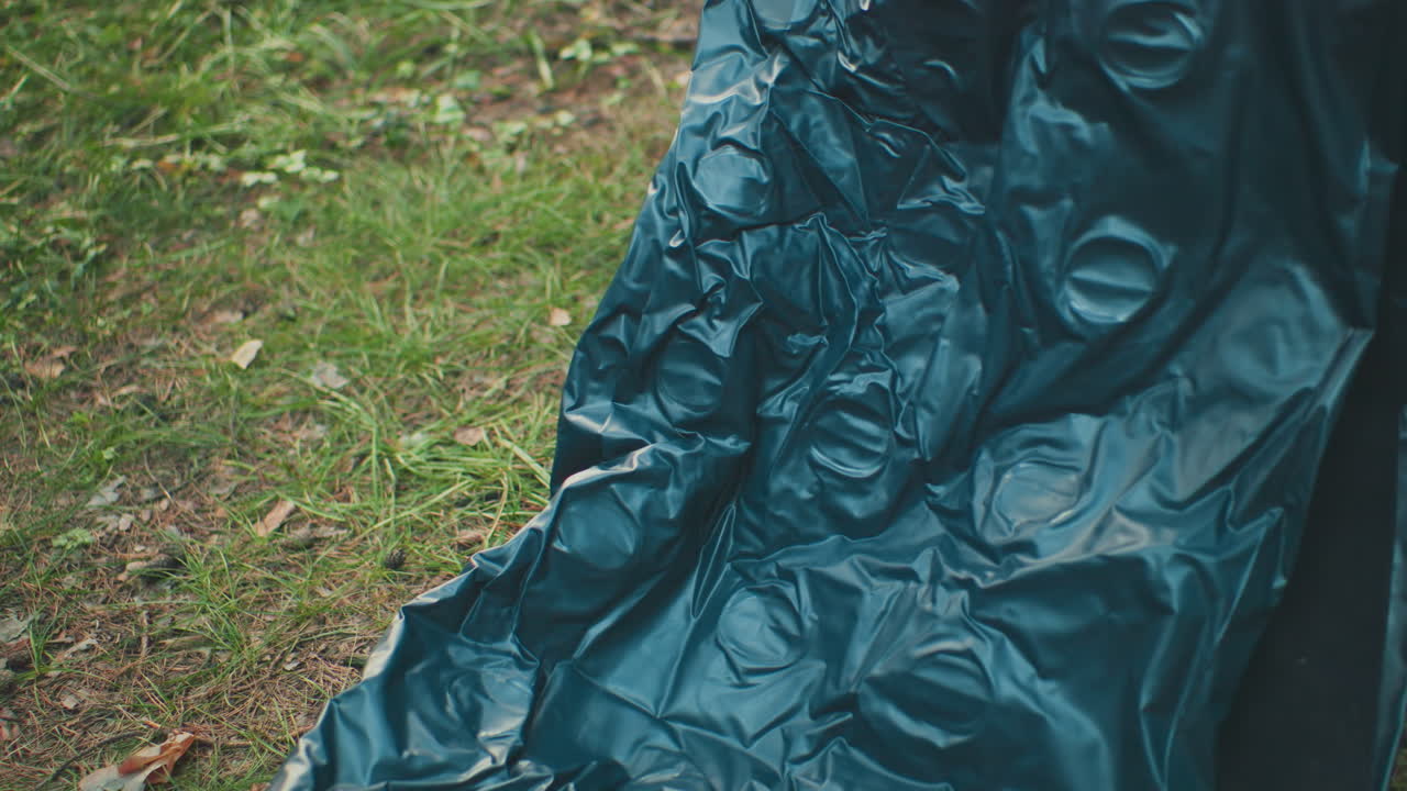 Close up of crinkled tent fabric as it is being opened on grassy ground with blurred surroundings, showing material texture and outdoor setup action in natural forest environment