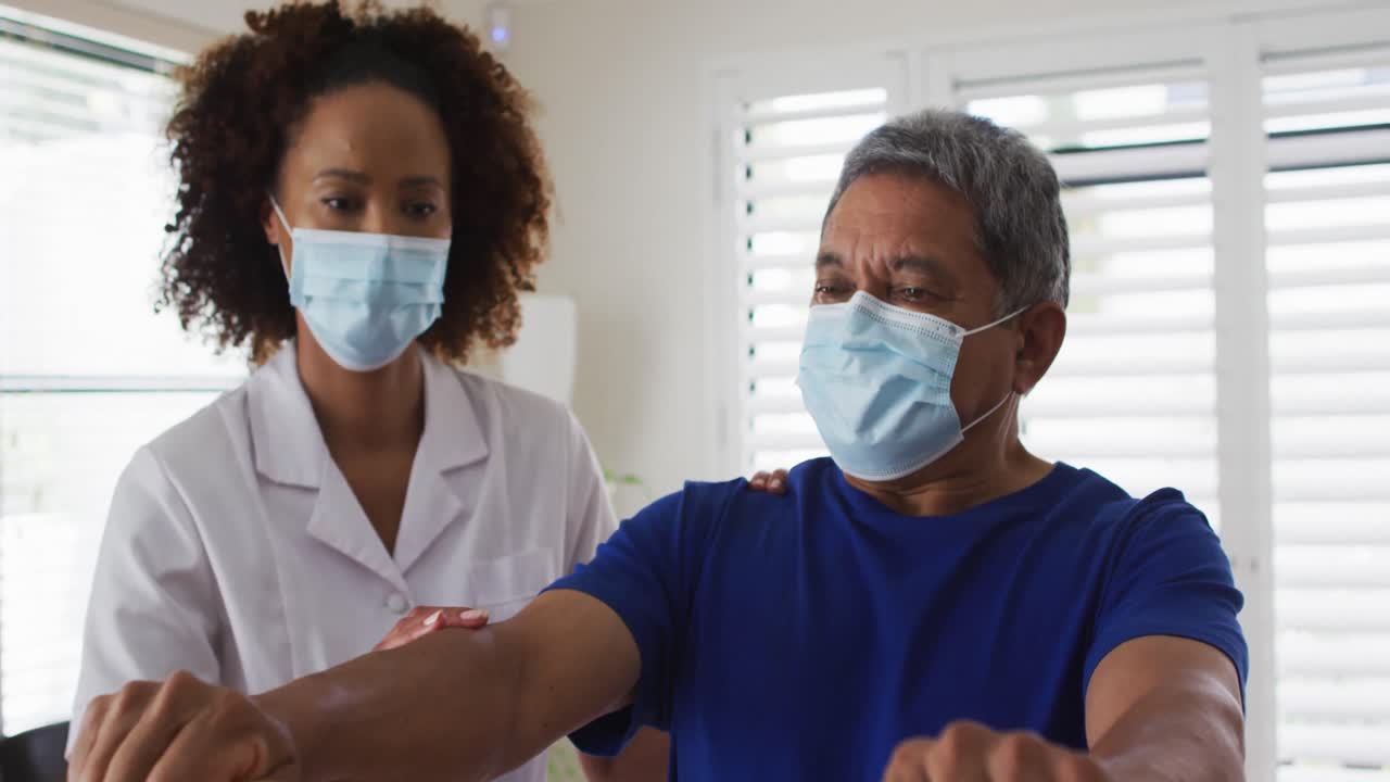 Mixed race female physiotherapist wearing mask helping senior exercise using exercise band