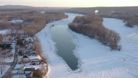 Aerial-forward-dolly-over-hudson-river-winter-with-a-partially-frozen-strip-of-water-beside-an-island,-foreground-shows-shoreline-houses-background-a-plume-of-steam-from-a-power-plant-and-mountains