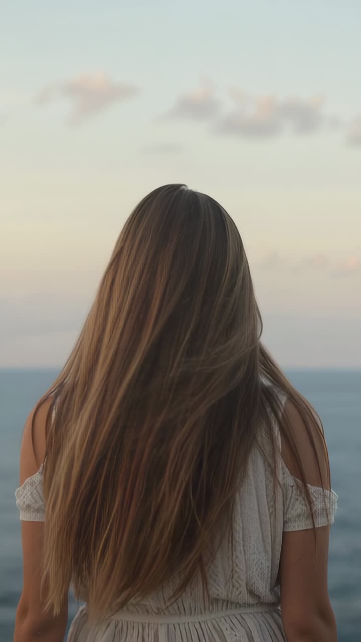 Vertical video: Breeze picking up, causing brown hair lifting and swaying behind woman at coastline