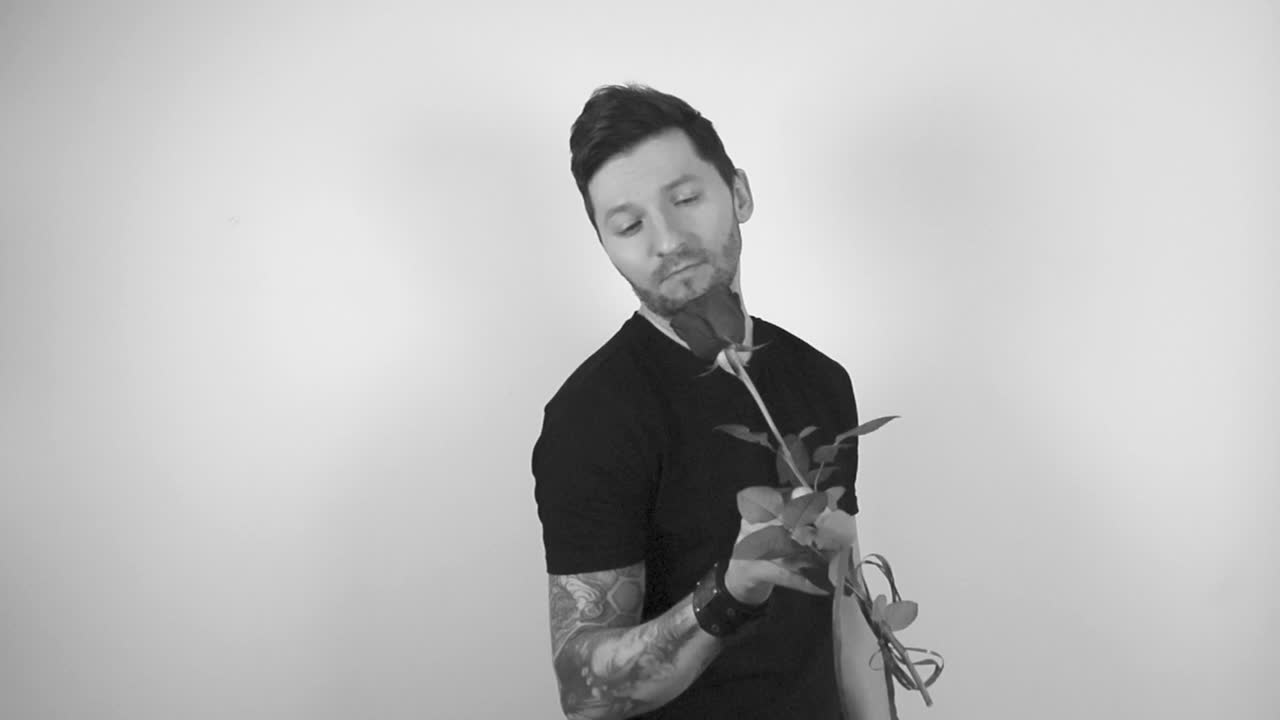 Grayscale footage of a tattooed man with short hair and trimmed beard looking at a blossoming rose flower, smelling it and offering in front of him to the camera or the viewer in slow motion.