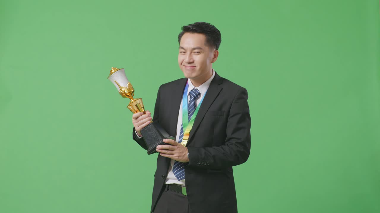 슈트와 타이를 입은 아시아 사업가, 금메달과 트로피를 들고 골을 치며, 스튜디오의 녹색 스크린 배경에서 첫 번째 우승자로 우승을 축하합니다.