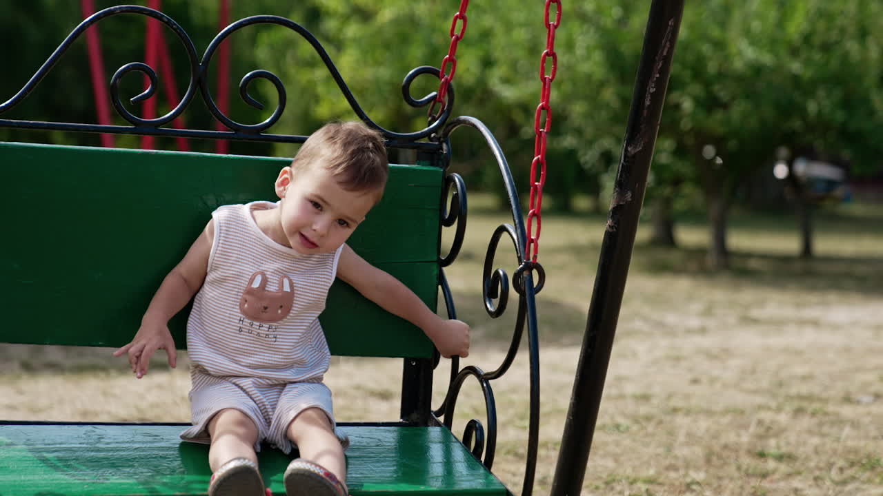 Adorable toddler boy swinging on the green swing in the park. Happy time outdoors in summer.