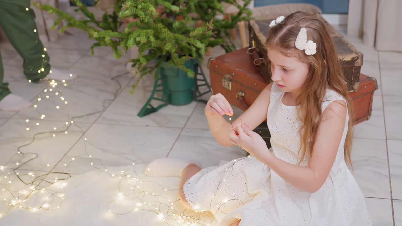 Young girl in white dress sitting on floor and carefully arranging tangled Christmas lights. Background features a Christmas tree, boxes, and a cozy holiday setting