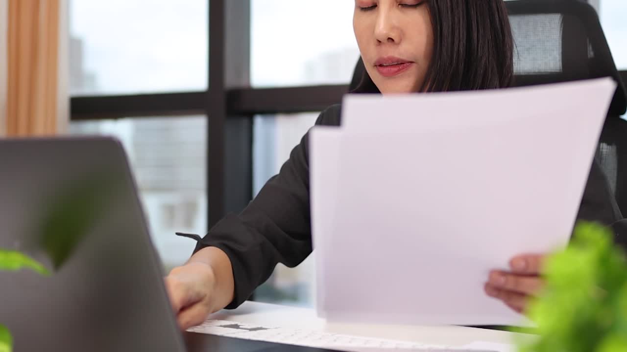 Una professionista si concentra sulla revisione di documenti mentre lavora al laptop in un ufficio luminoso.
