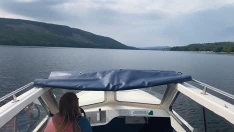 woman with blonde hair driving a small white motor boat down a vast lake in coniston - cumbria, uk