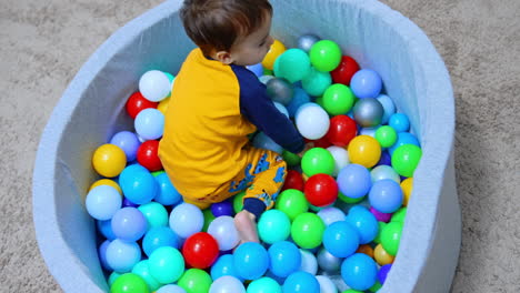 kid in yellow pajama climbs into the dry pool. baby boy sits into the colorful balls. top view.