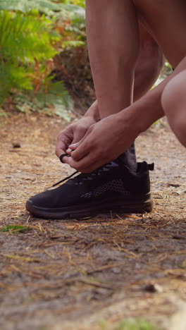 vertical video close up of man tying laces on training shoe before exercising running along track through forest shot in real time