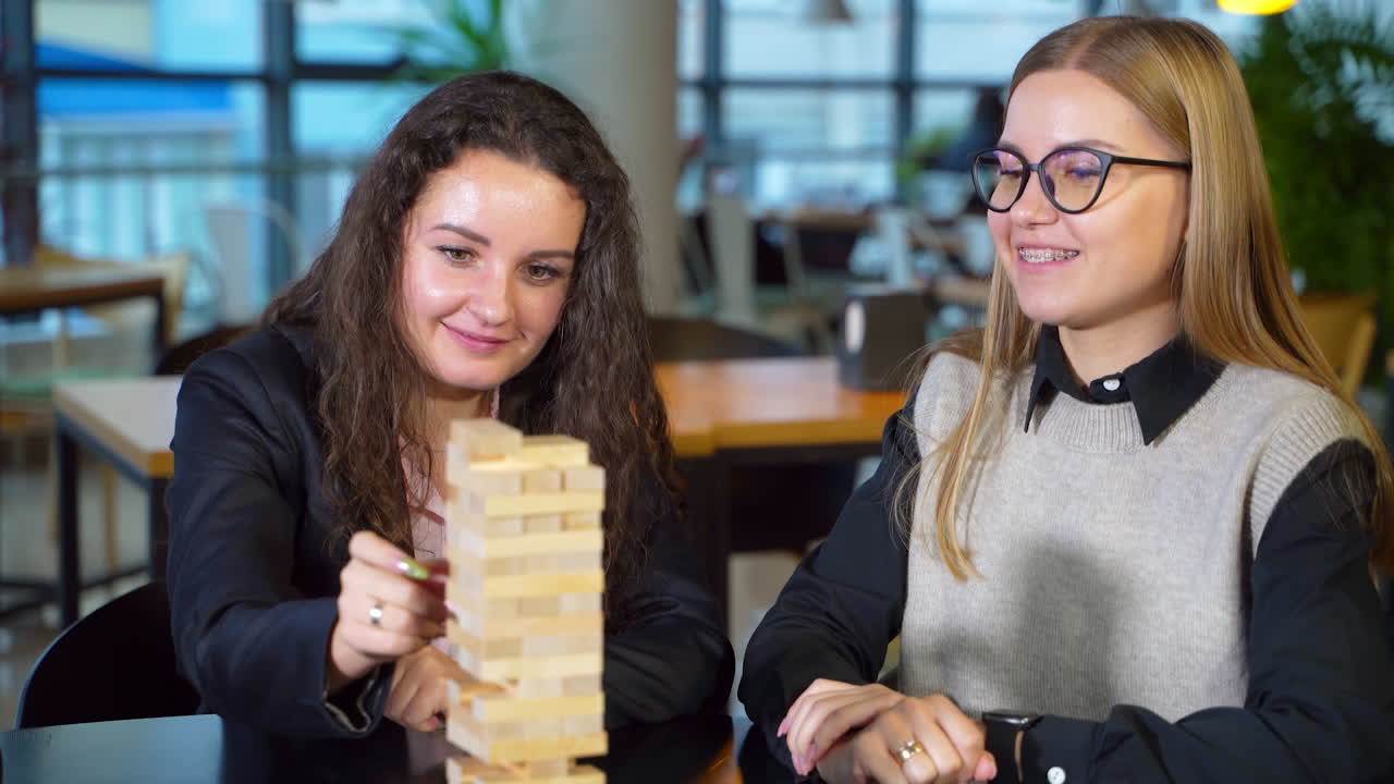 Young woman with long dark curly hair pulls a wooden brick slowly. Lady puts a piece on the pile carefully. Girl sitting next smiling cheerfully.