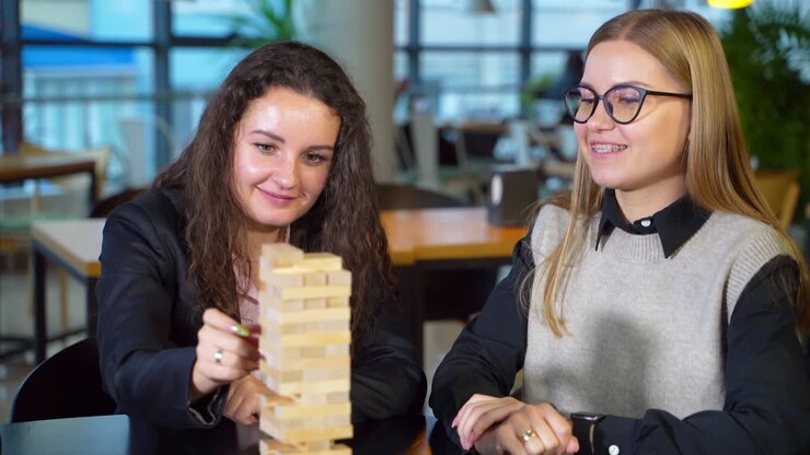 Young woman with long dark curly hair pulls a wooden brick slowly. Lady puts a piece on the pile carefully. Girl sitting next smiling cheerfully.