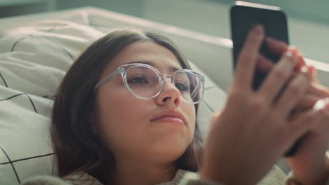 Zoom-in-of-caucasian-teenage-girl-browsing-phone-while-lying-on-bed-on-her-back